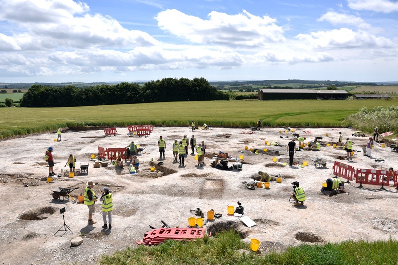 A wide shot of the excavation site showing pits, students working and farm fields in the background
