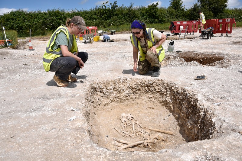 Two female student archaeologists crouching over a pit containing human bones.