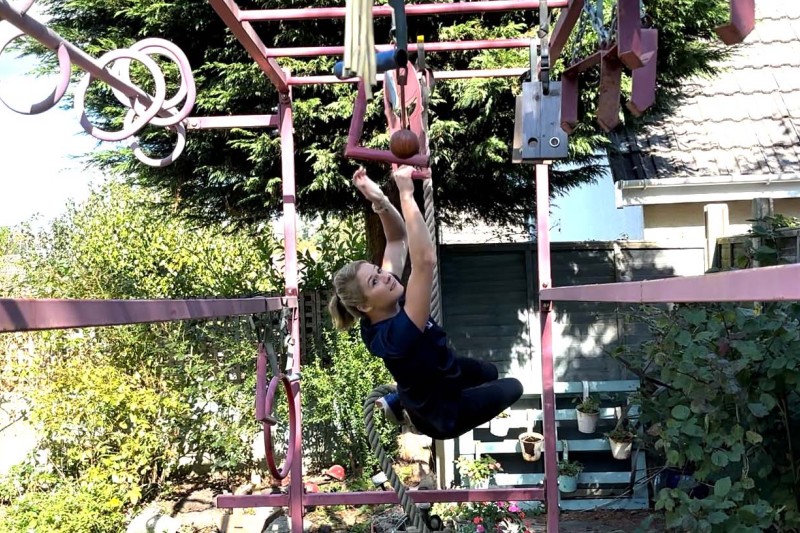 Becky on monkey bars, grabbing a rope with her legs and reaching for a handle with her hand