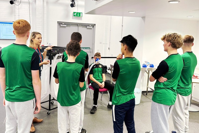 Dr Becky Neal talking to a group of young male students, one of them is sitting on a chair wearing device to measure his breathing