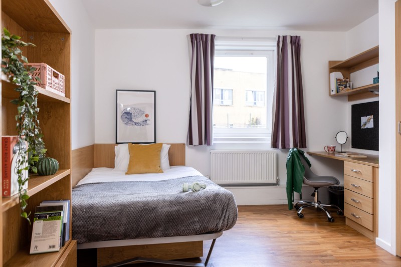 A 3/4 sized bed to the left of the room and bookcase. To the right there is a desk, desk chair, shelving and pinboard with a window in the centre of the image