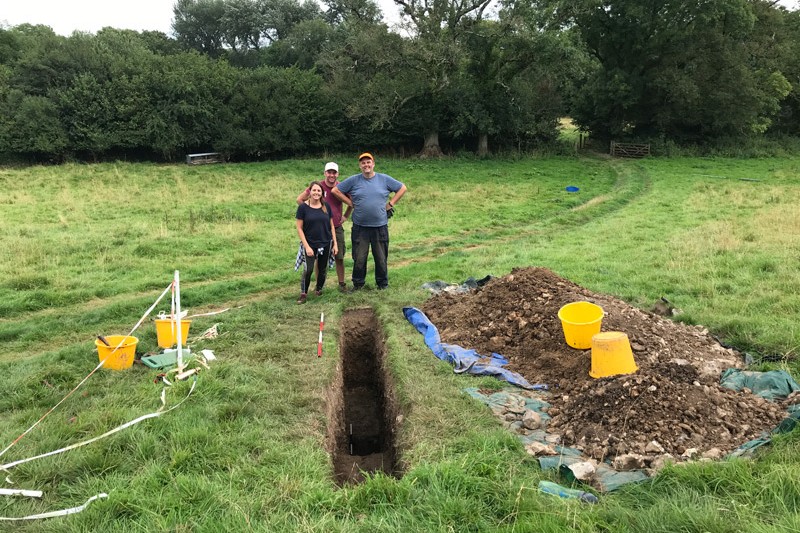 A woman and two men standing at the end of a rectangular trench in a grassy field, next to the trench is a big pile of earth which they had excavated