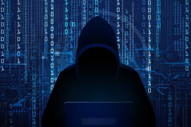 A hooded silhouetted figure on a laptop in front of computer code