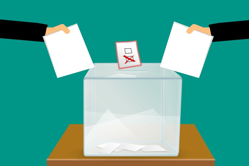 A cartoon image of two hands holding pieces of paper above a ballot box