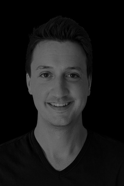 This black and white head shot shows Sam Taylor smiling to the camera. there is a balck background and he wears a black V-neck t-shirt. 