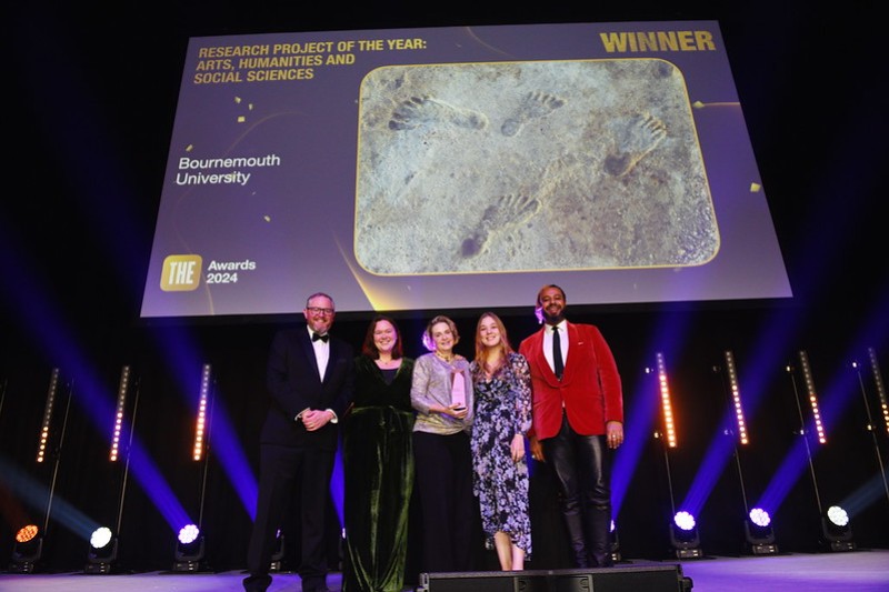 Three women and two men on a stage beneath a big screen showing the ancient footprints that were studied in this research. The lady in the middle of the group is holding the award they have just collected. 