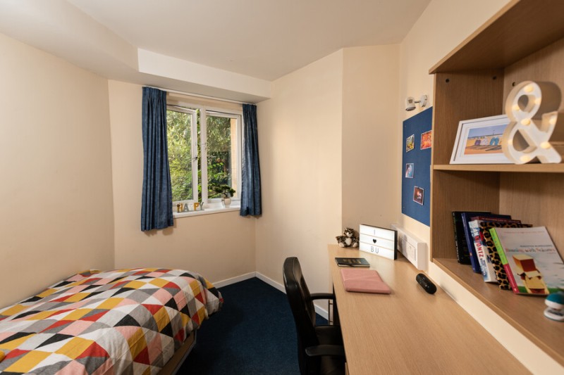 A single bed and window to the left of the image and desk, shelving and pinboard to the right