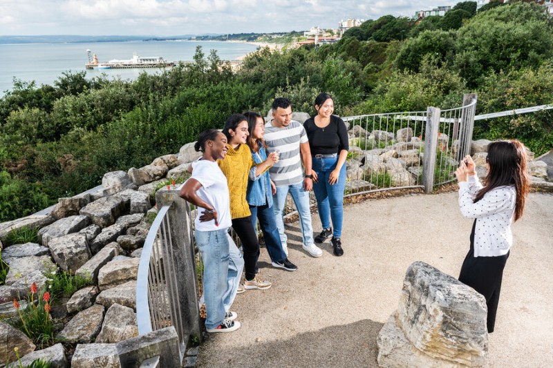 A photo of students posing for a picture at the beach. Students are from UK and international countries, they stand in a semi-circle and post the camera on the cliff top. The sea and Bournemouth pier are in the background of the image. 