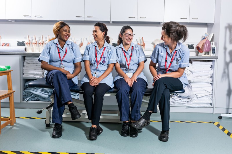 Four nursing students are pictured in their uniforms, all sat on one of the ward beds laughing as they take a break in the simulation wards.